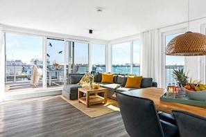 Un salon moderne avec un canapé, une table basse et une table à manger, de grandes fenêtres offrant une vue sur la ville et un luminaire suspendu.