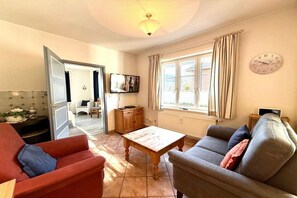 A living room with a sofa, a coffee table, a television, and a window with curtains. - House 94 - Apt. Ground Floor (Oldsum)