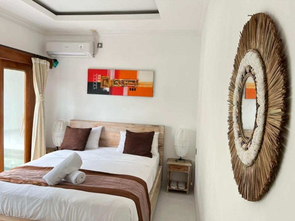 A hotel room with a bed, a wall-mounted air conditioner, a framed artwork, and a decorative wall piece.