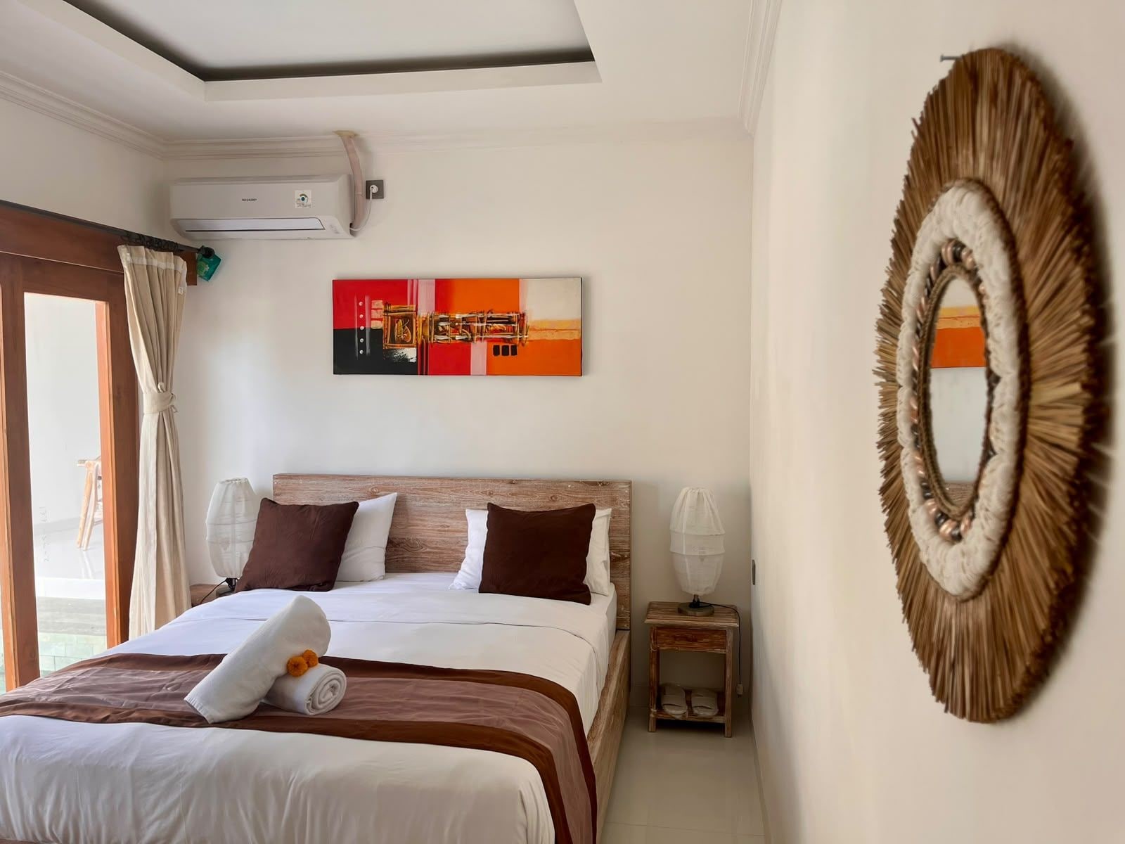 A hotel room with a bed, a wall-mounted air conditioner, a painting, a decorative mirror, and a window with curtains.