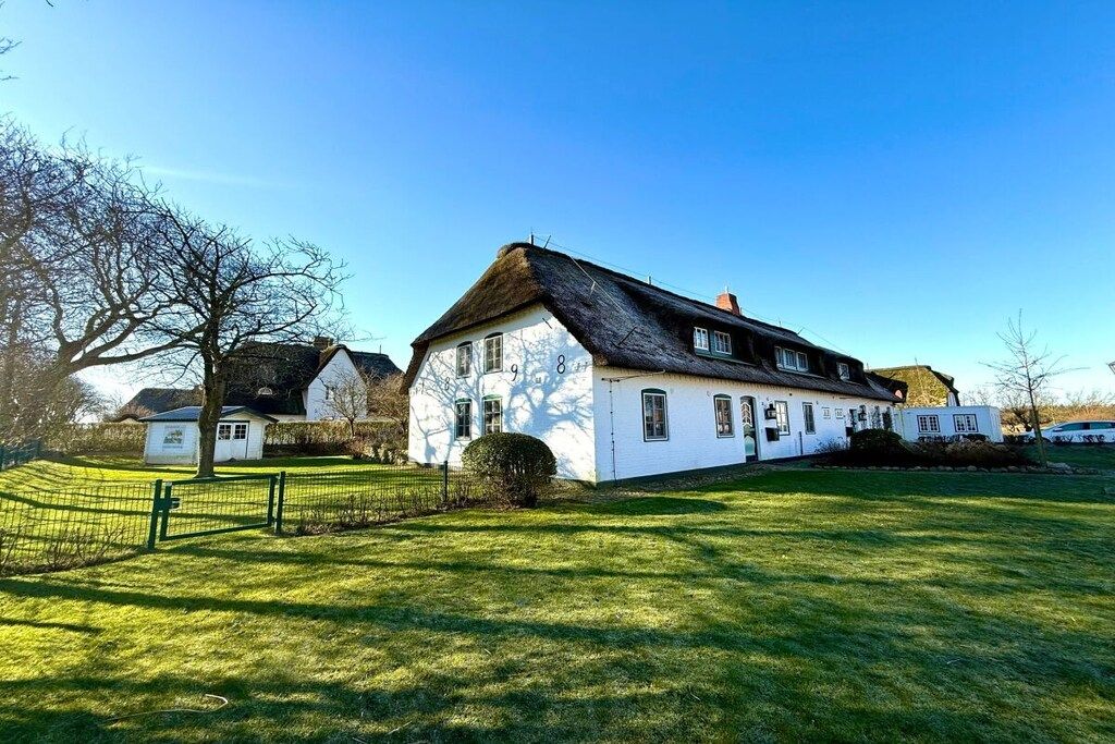 A white house with a thatched roof, surrounded by a green lawn and a fence.