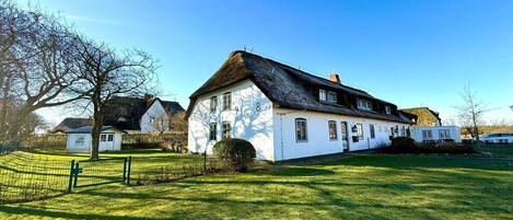 A white house with a thatched roof, surrounded by a green lawn and a fence.
