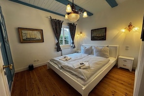 A bedroom with a white bed, a wooden floor, a window with curtains, and a chandelier. - Det Danske Hus - Apartment Starboard (Dunsum)
