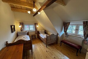 Quarto com duas camas de madeira, um banco, uma mesa e janela com cortinas.