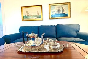 A room with a blue sofa, a wooden table with a silver tea set, and two framed maritime paintings on the wall. - Det Danske Hus - apartment on the port side (Dunsum)