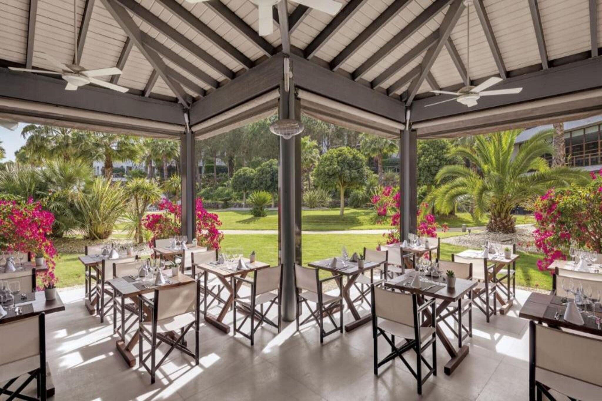 Un área de comedor al aire libre con mesas y sillas, rodeada por un jardín con diversas plantas y flores.