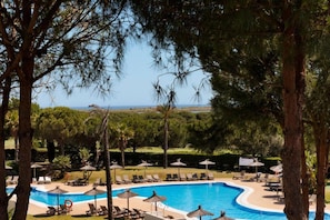 A pool area with lounge chairs and umbrellas, surrounded by trees and a view of the sea. - El Rompido Resort- The Hotel by Sunday- Standard Double Room (El Rompido)