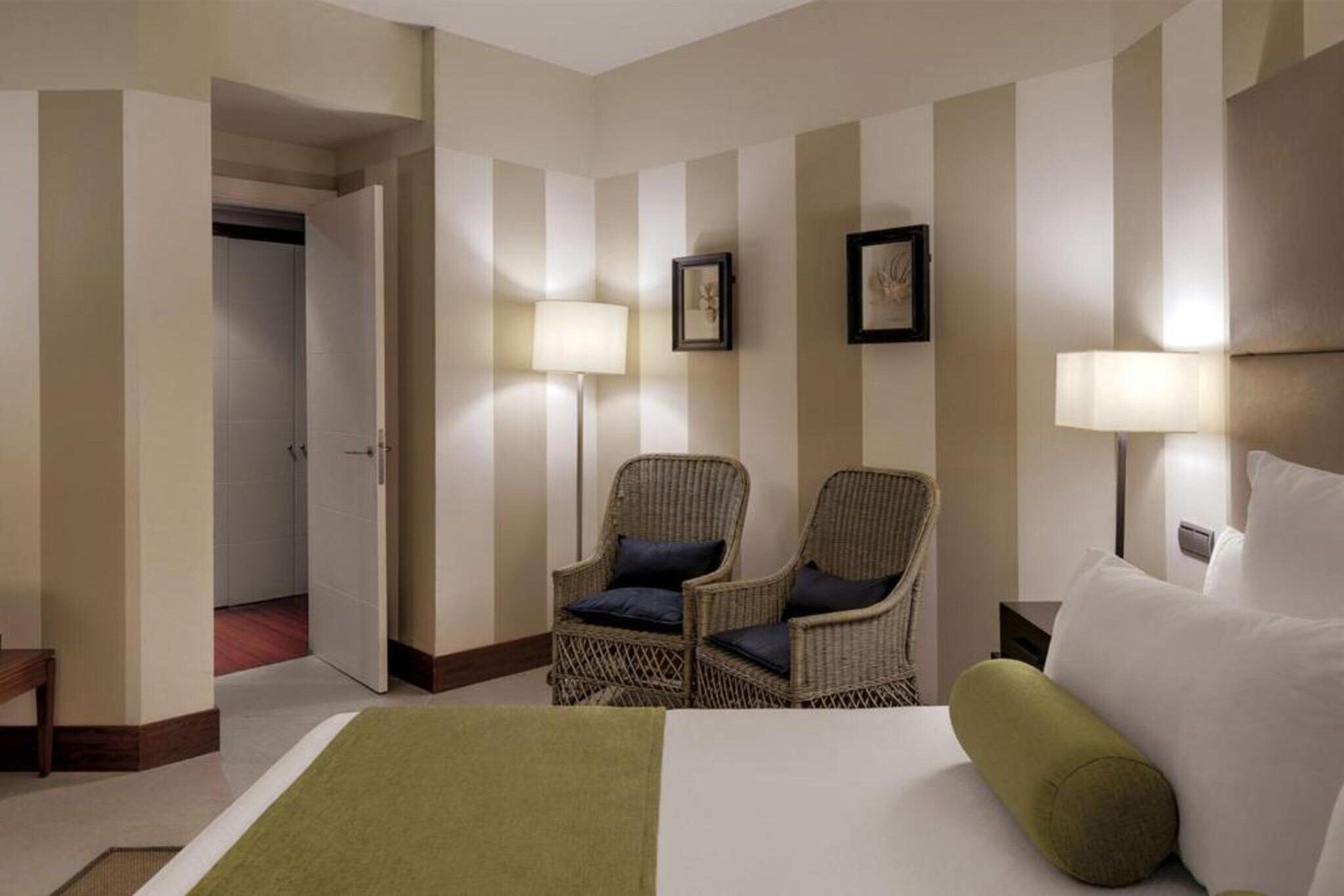 Una habitación de hotel con una cama, dos sillones de mimbre, una mesita de noche y dos lámparas de pared.