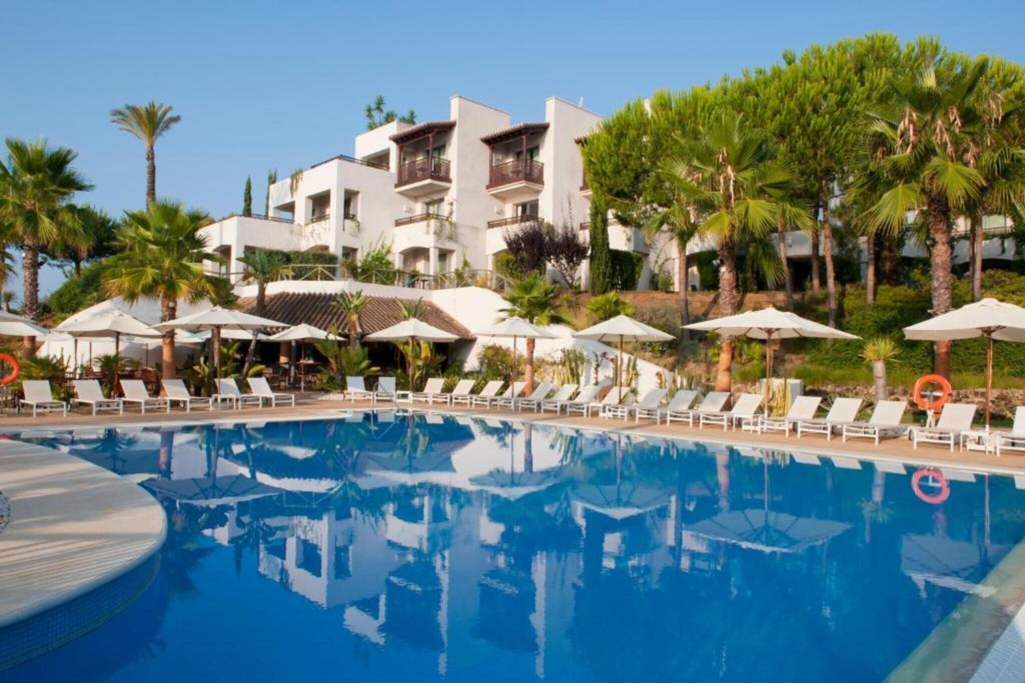 A swimming pool with lounge chairs and umbrellas, surrounded by palm trees and a building with balconies.