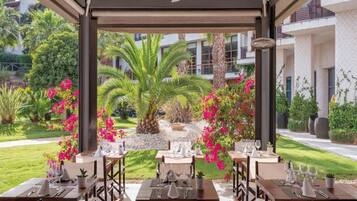 Un área de comedor al aire libre con mesas dispuestas para dos, rodeada de follaje exuberante y plantas florecientes.