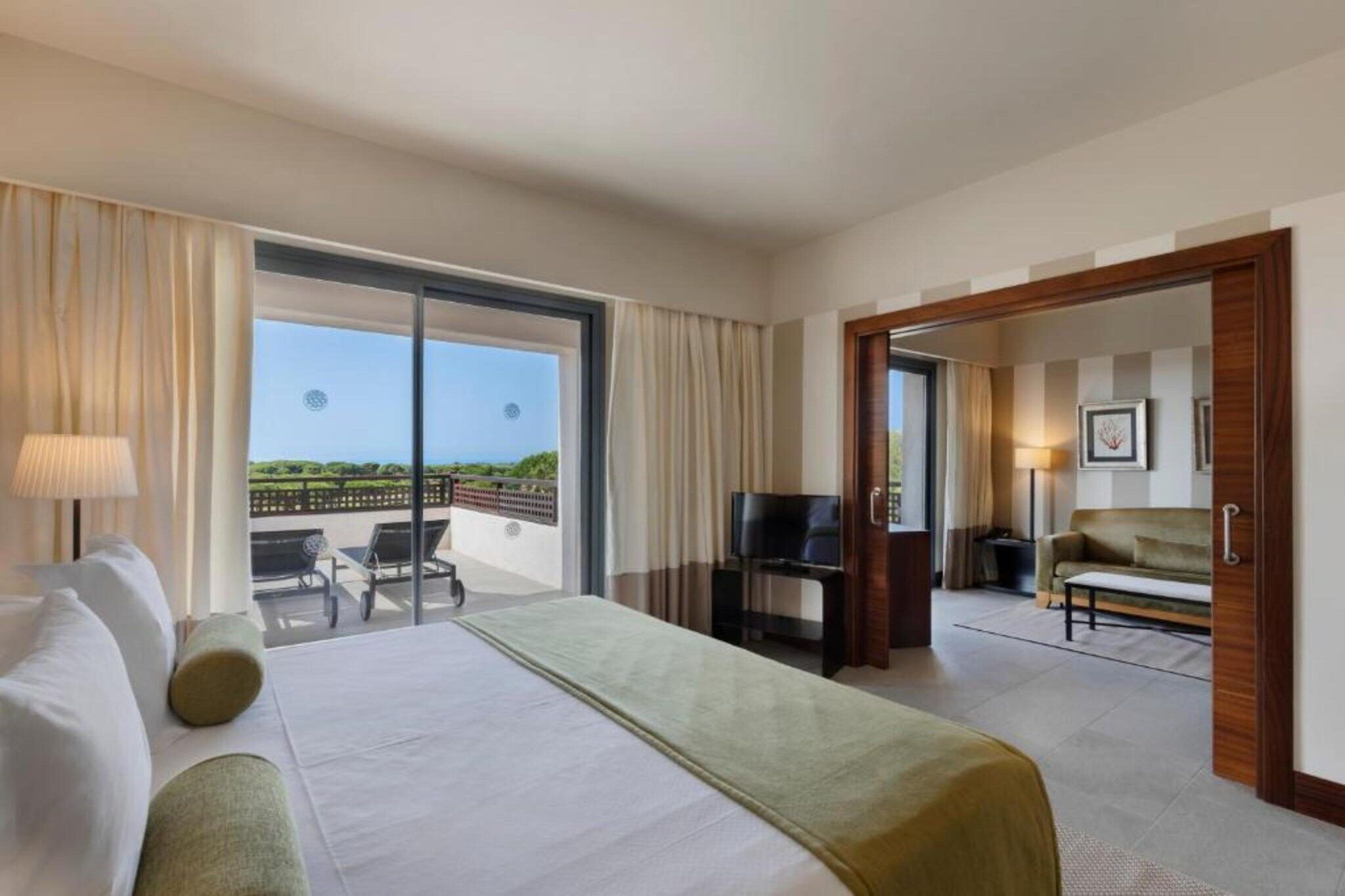Habitación de hotel con cama, televisión, sofá, silla y balcón con vistas.