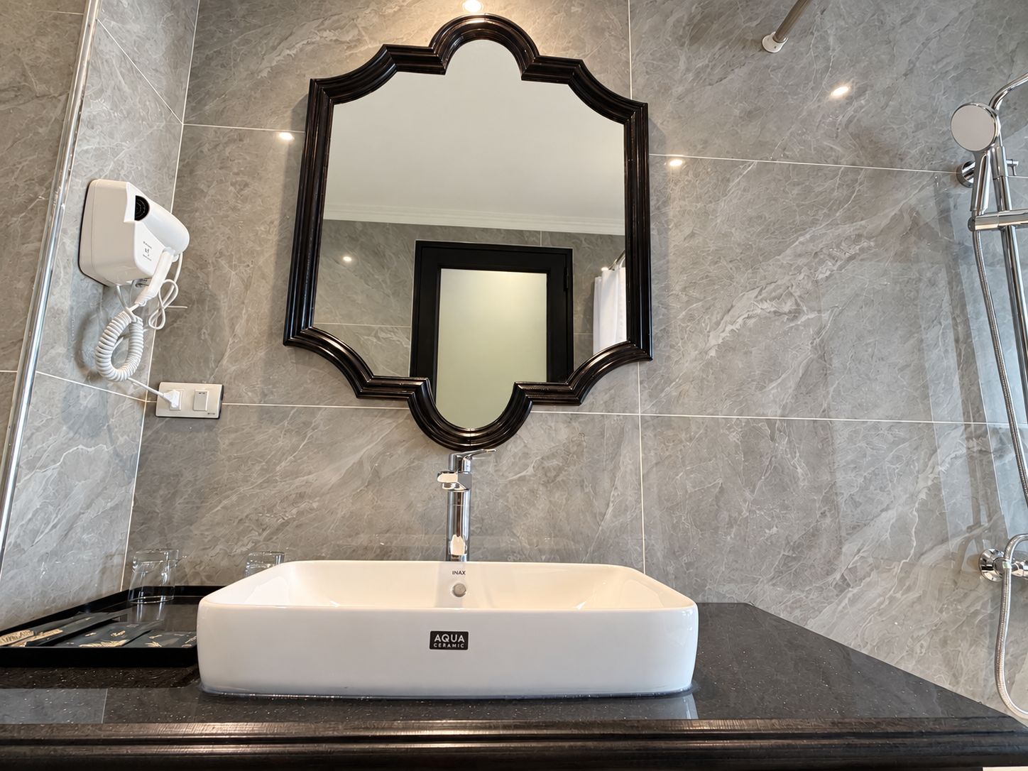 Casa de banho com lavatório de cerâmica branca, espelho com moldura preta e secador de cabelo portátil na parede.