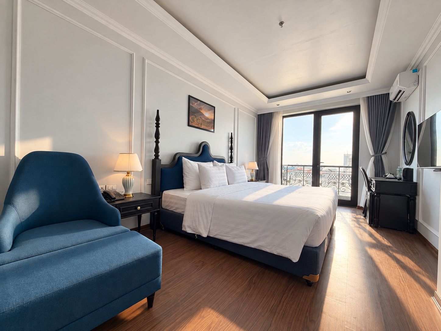 A hotel room with a large bed, a blue armchair, a desk with a lamp, and a view of the city through the window.