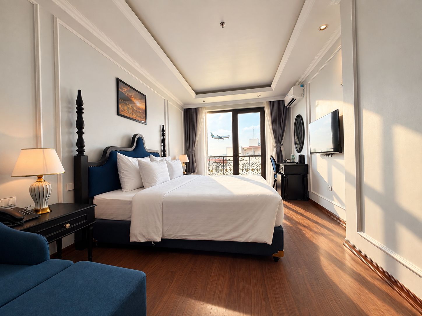 A hotel room with a bed, a desk with a lamp, a TV, and a view of the sky.