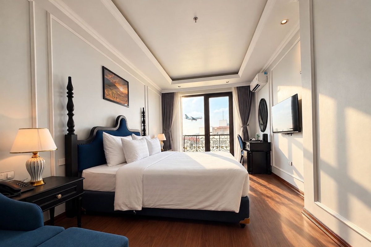 Quarto de hotel com cama, escrivaninha com luminária, TV e vista do céu.