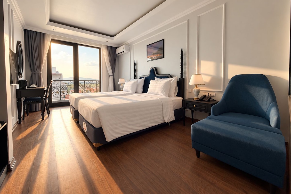 Quarto de hotel com uma cama grande, uma escrivaninha com cadeira, uma poltrona azul e uma varanda com vista.