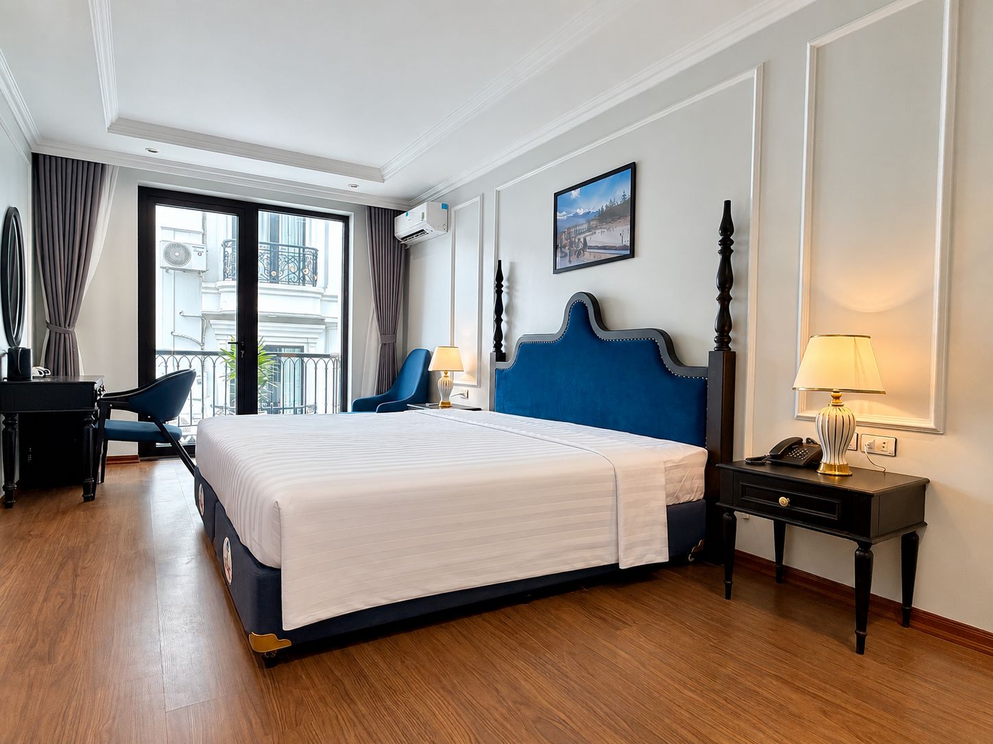 Quarto de hotel com uma cama grande, uma escrivaninha, uma cadeira, mesa de cabeceira com abajur e janela com cortinas.