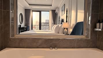 A hotel room with a large bed, a sitting area, a bathroom with a bathtub, and a city view through the window.
