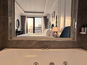 Quarto de hotel com cama grande, área de estar, banheiro com banheira e vista para a cidade pela janela.