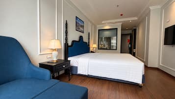 Ein Hotelzimmer mit einem Bett, einem blauen Sessel, einem Nachttisch mit Lampe, einem großen Spiegel und einem an der Wand befestigten Fernseher.