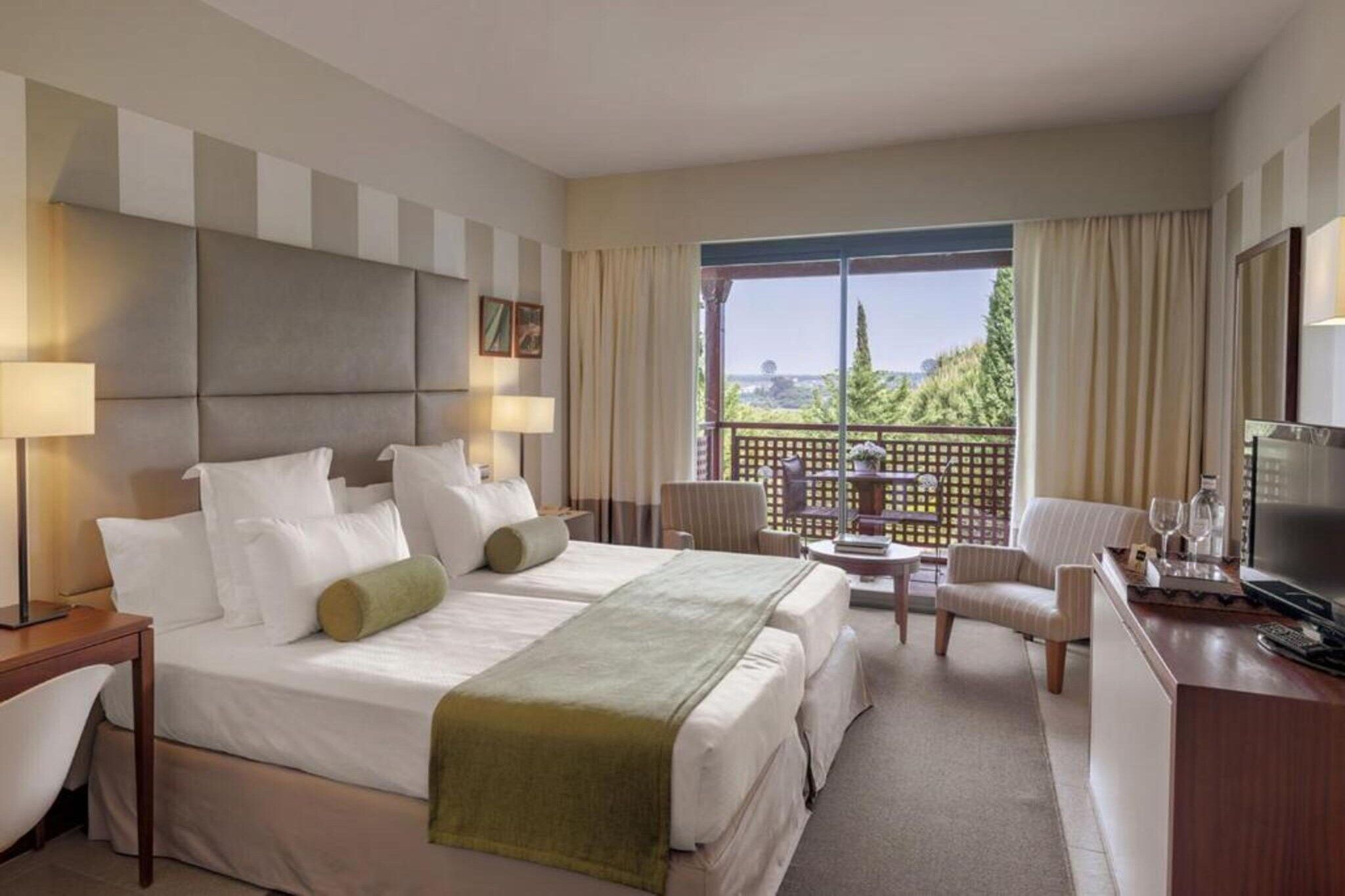 A hotel room with a large bed, a desk with a TV, a chair, and a balcony with a view.