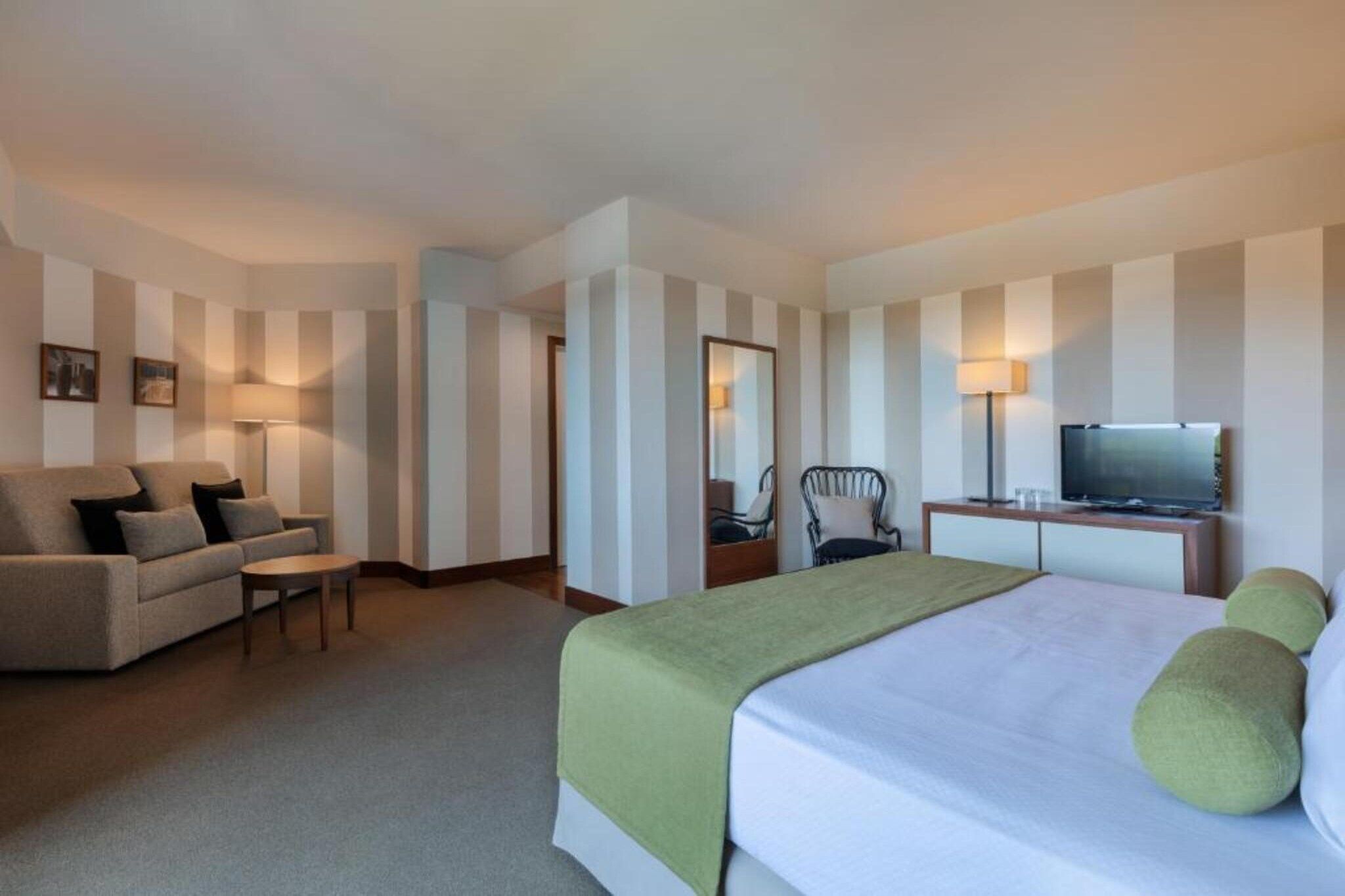 Una habitación de hotel con cama, sofá, sillón, escritorio con televisor y espejo.
