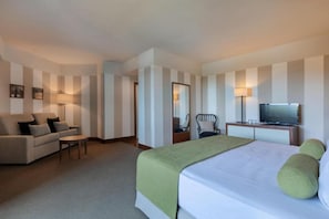 A hotel room with a bed, a sofa, a chair, a desk with a television, and a mirror.