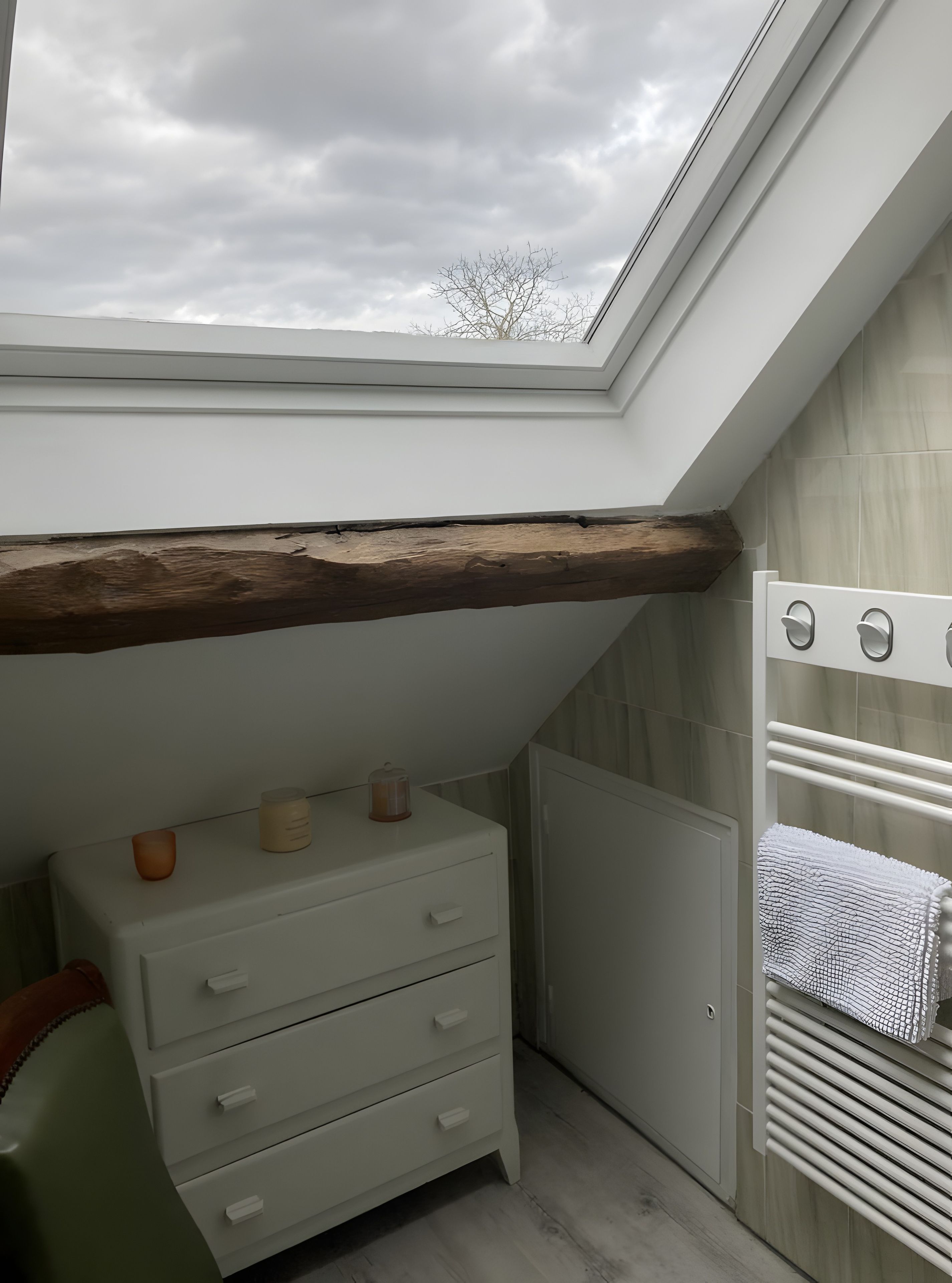 Een slaapkamer met dakraam, een houten balk, een radiator en een witte dressoir met laden.