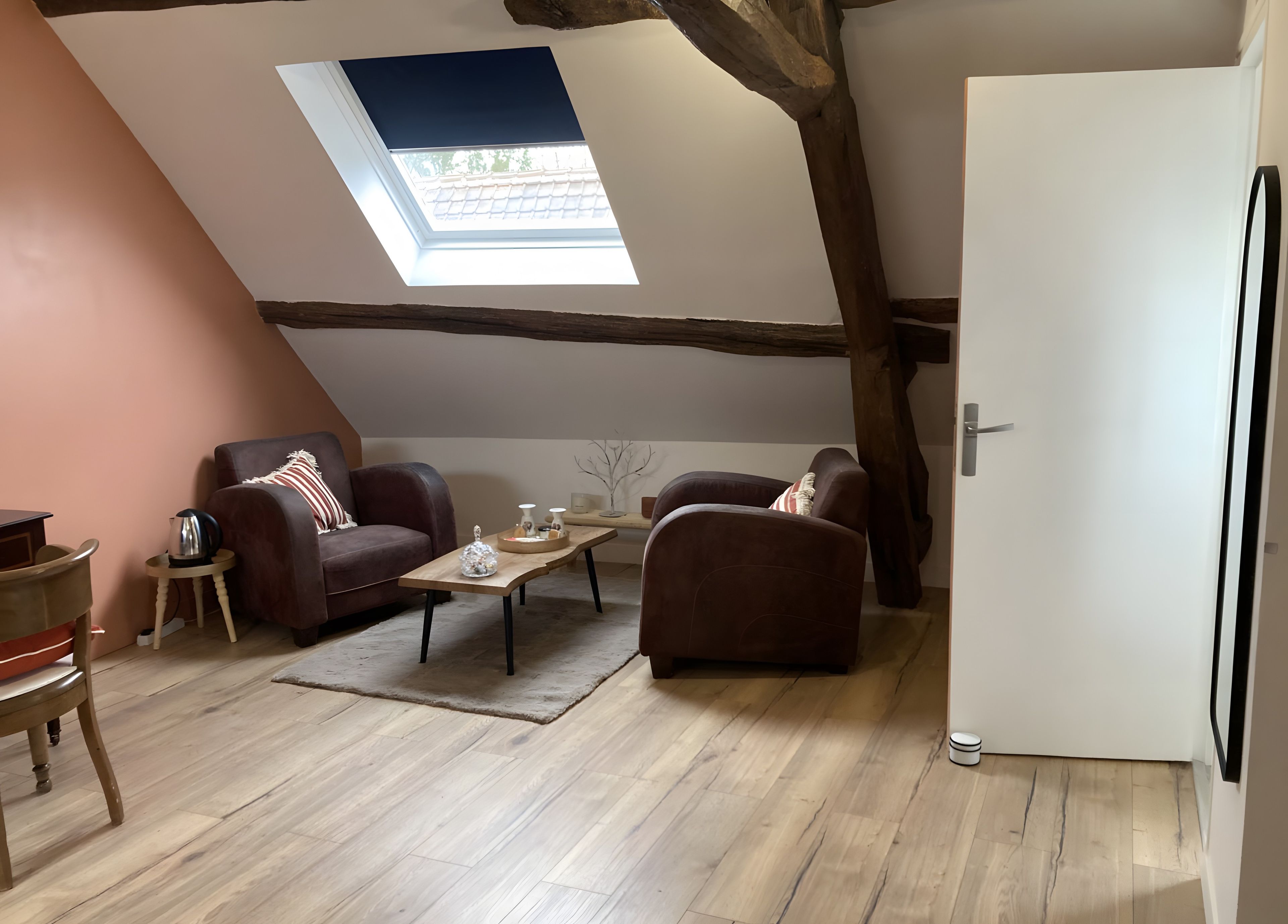 Een loft-achtige kamer met een schuine kap, dakraam, houten balken, een bank, een salontafel en een deur.