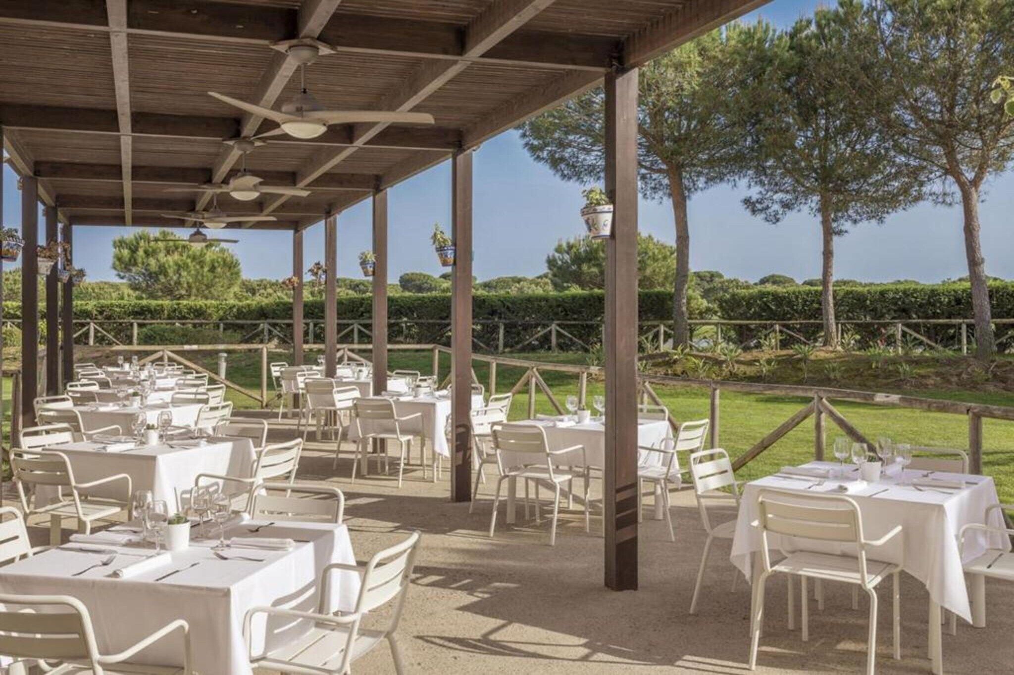 Un área de comedor exterior con mesas y sillas, cubierta por una estructura de madera con ventilador de techo.