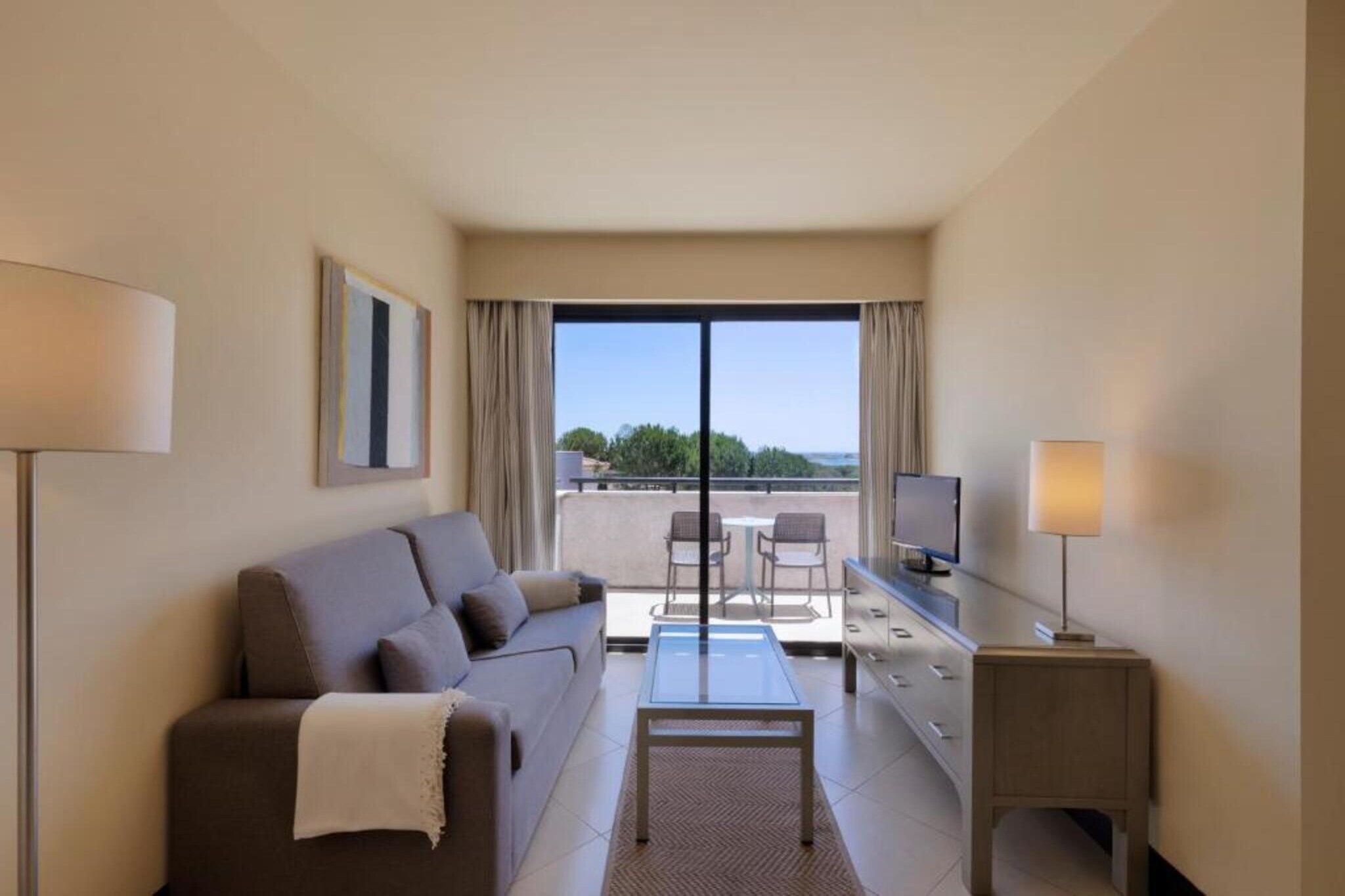A hotel room with a sofa, a coffee table, a TV, and a balcony with a view.