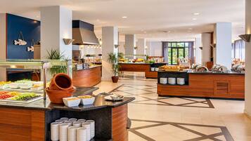 A hotel breakfast area with multiple food stations, a central serving counter, and a view of the outdoors through large windows.