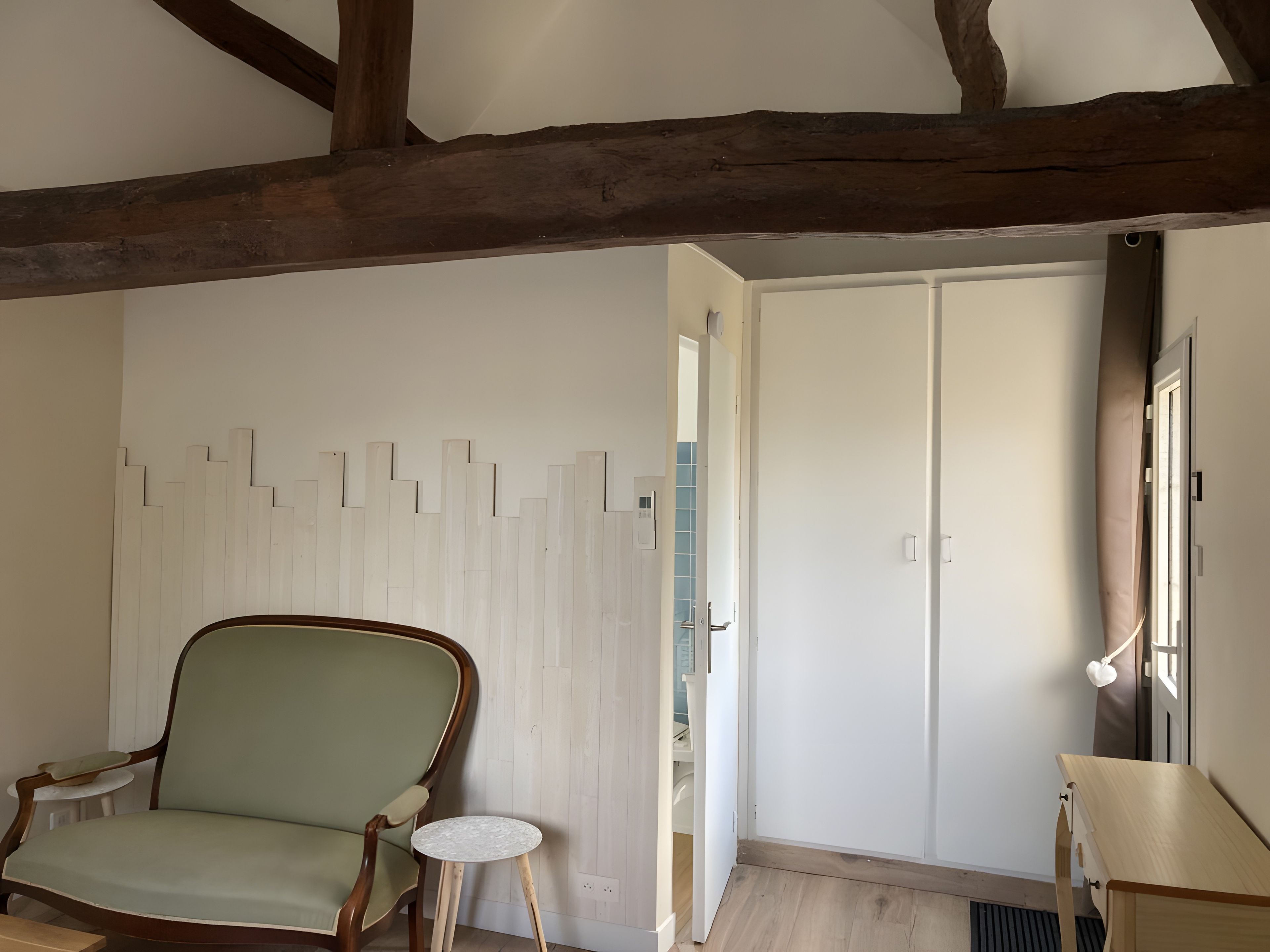 Een kamer met een houten plafond, een groen bekleden stoel, een klein rond tafeltje, een kledingkast en een raam met gordijnen.