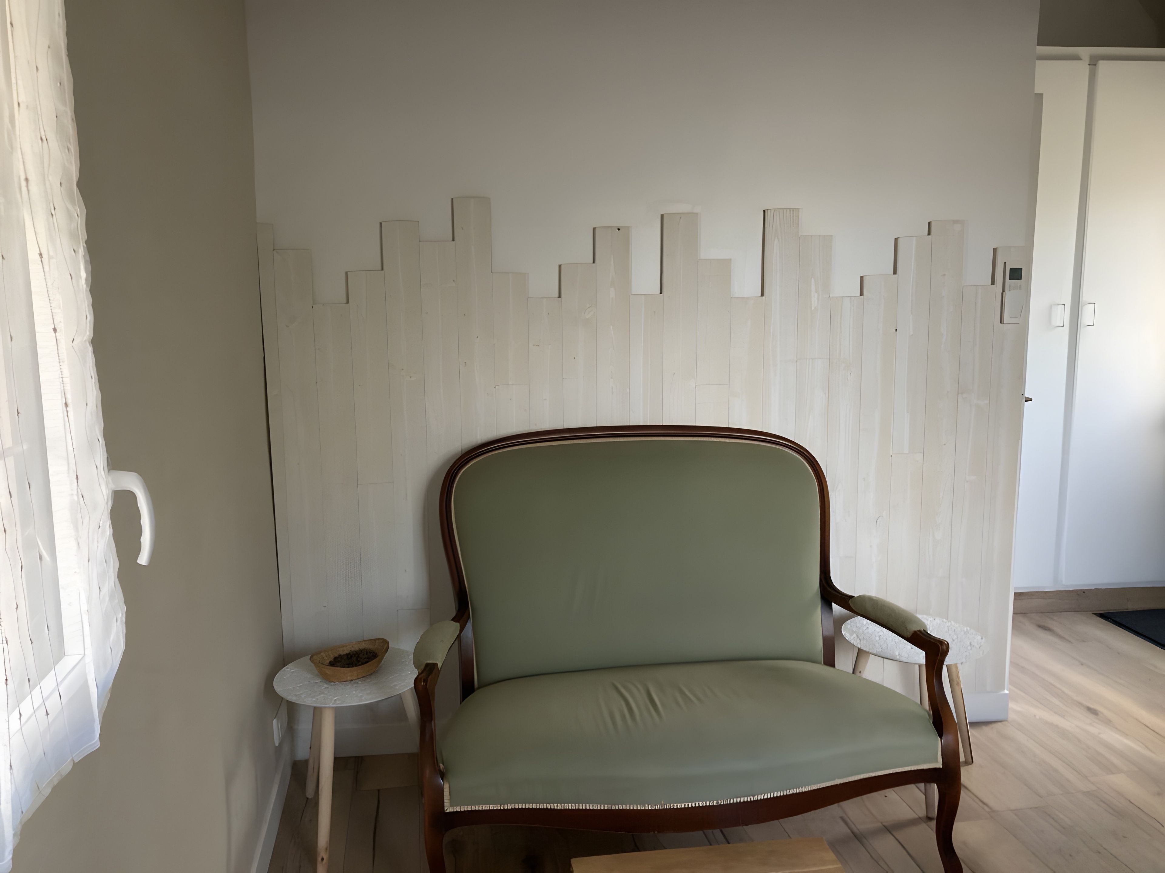 Una habitación con un sillón de madera, una mesita redonda y una pared blanca con diseño geométrico.