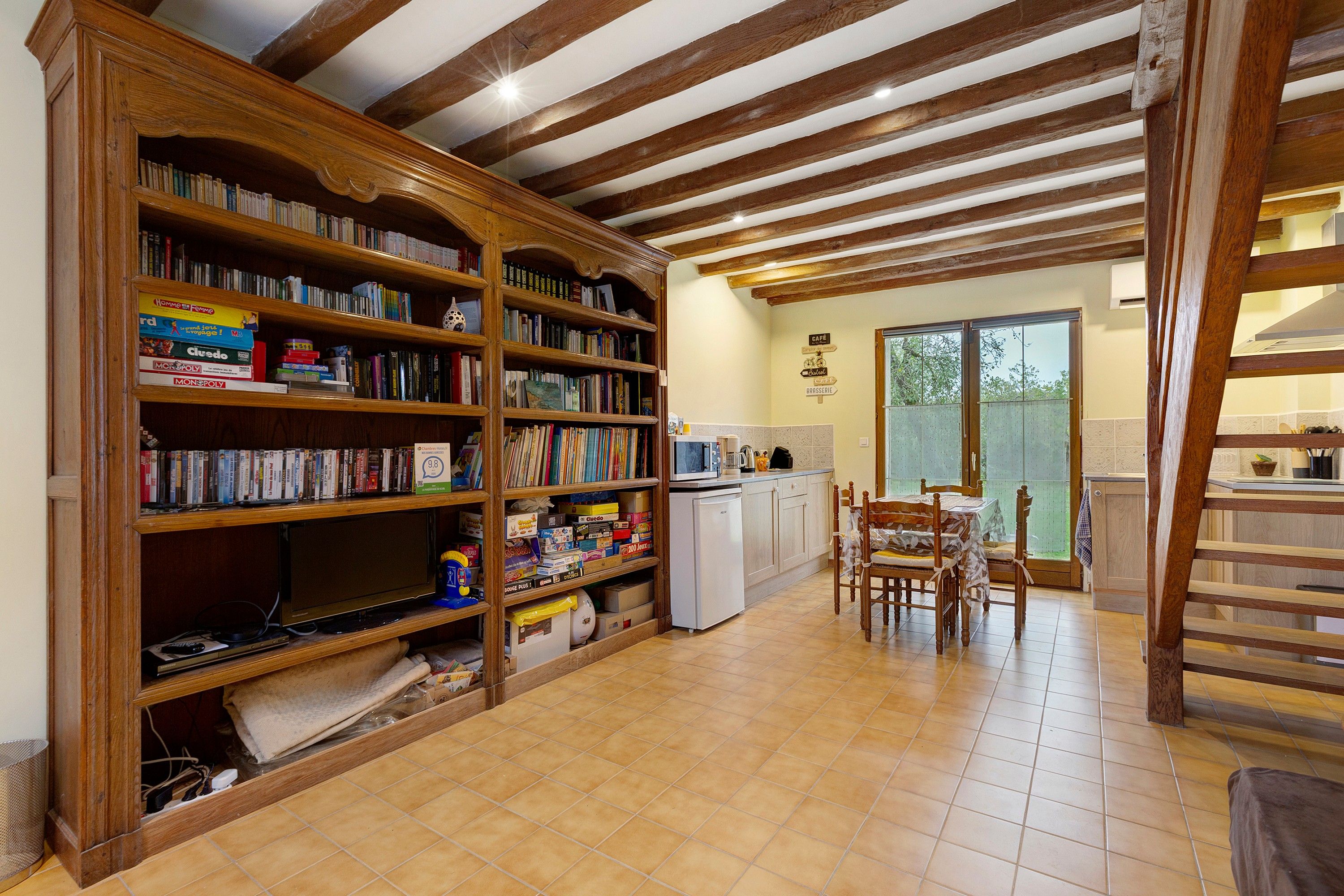 Een ruime kamer met een grote houten boekenkast vol boeken en gezelschapsspellen, een keukenhoek met een eettafel, en uitzicht door een glazen deur.