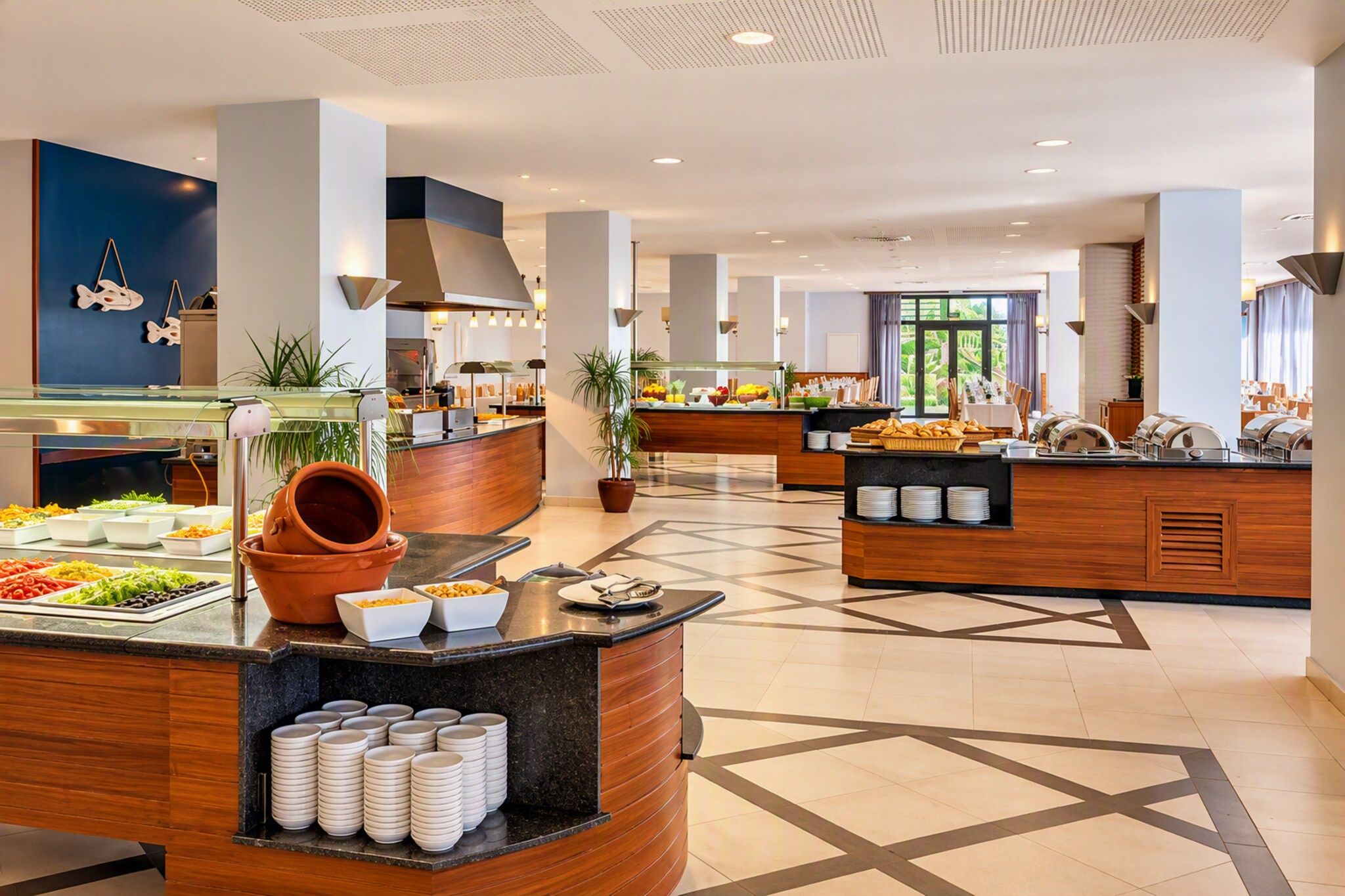 A hotel breakfast area with multiple food stations, a central serving counter, and a view of the outdoors through large windows.