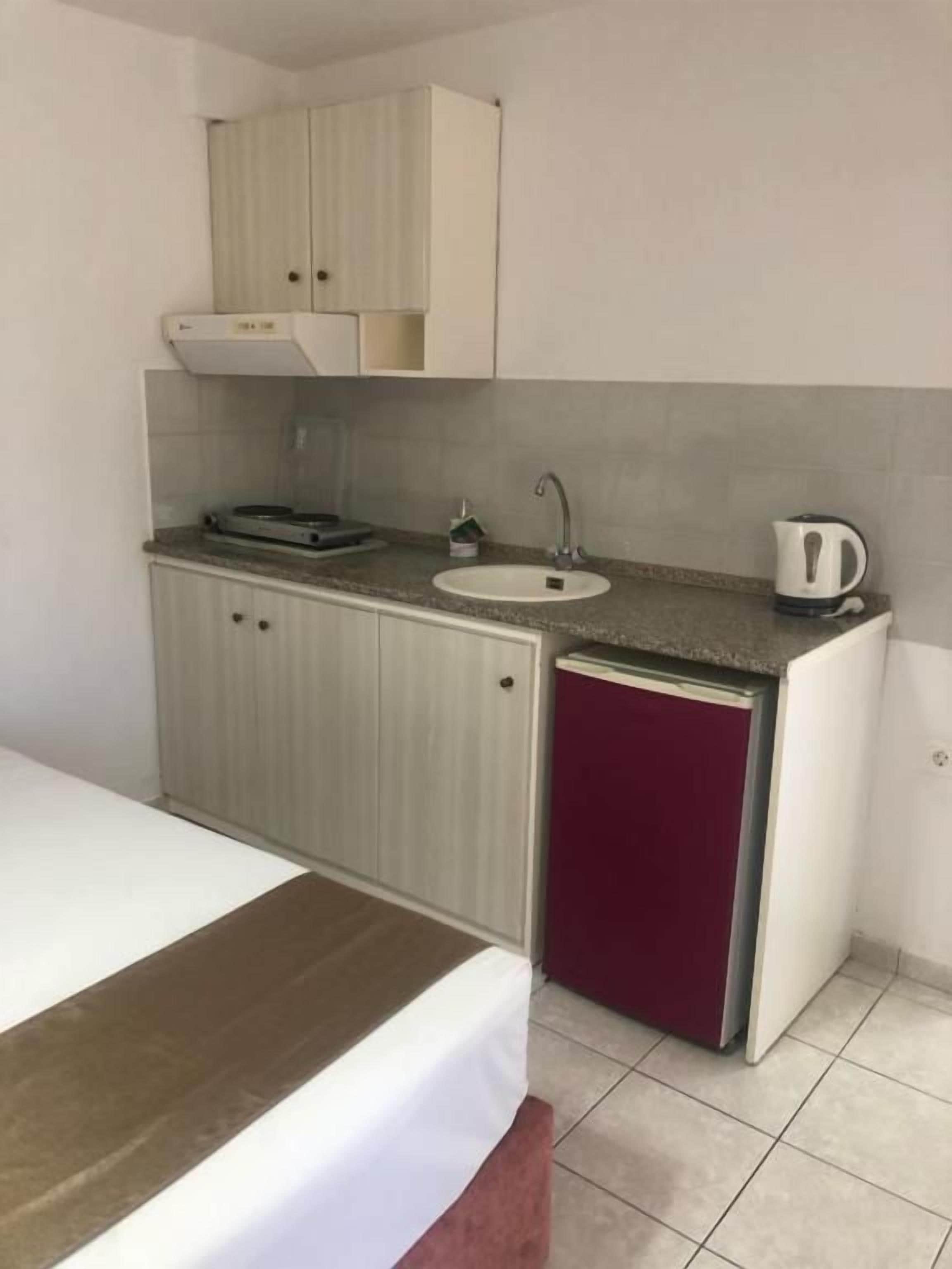 A compact hotel room with a bed, a kitchenette with a sink and a kettle, and a small cabinet above the sink.
