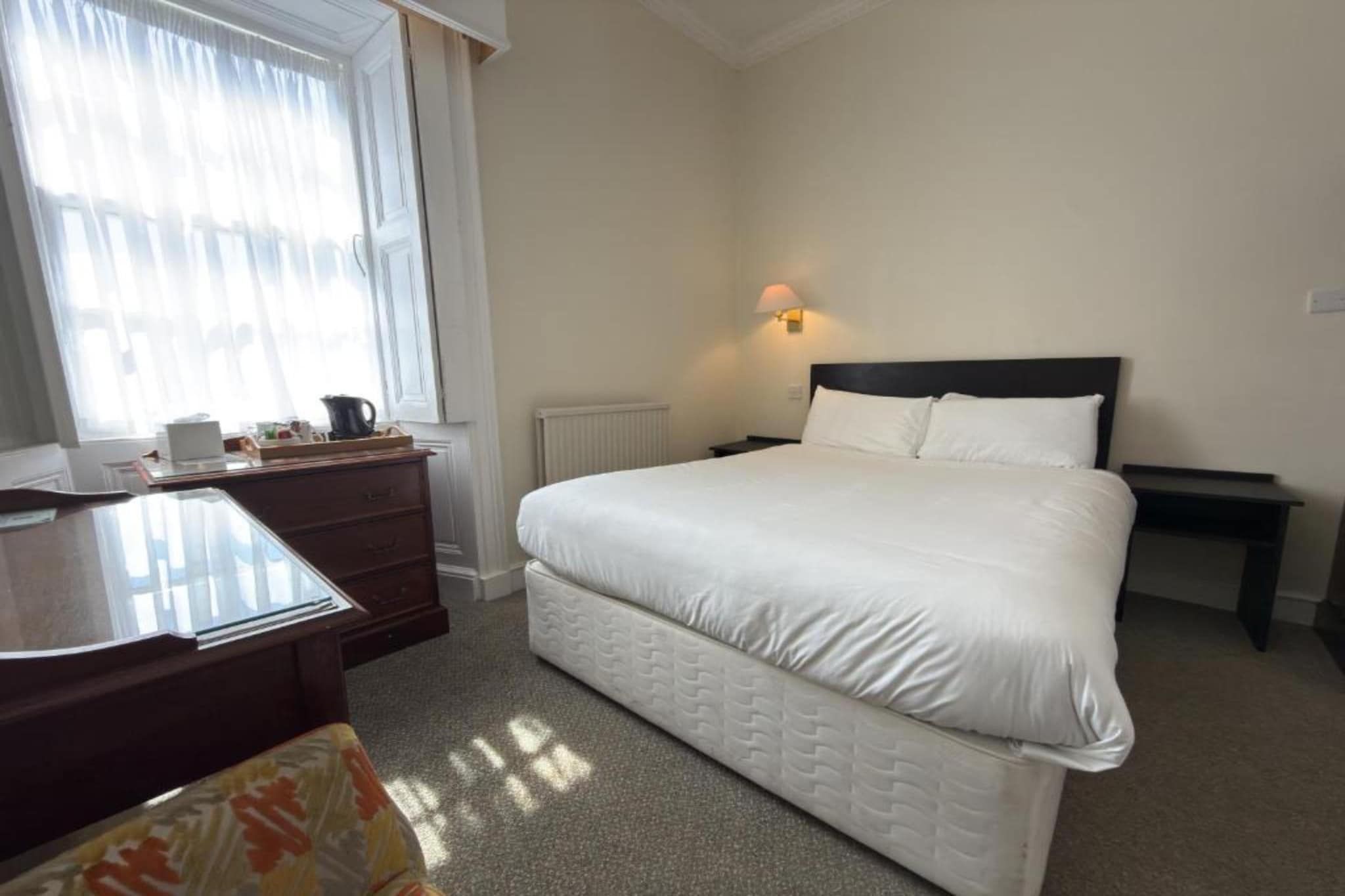 A hotel room with a bed, a desk, a chair, a radiator, and a window with curtains.