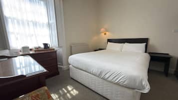 A hotel room with a bed, a desk, a chair, a radiator, and a window with curtains.