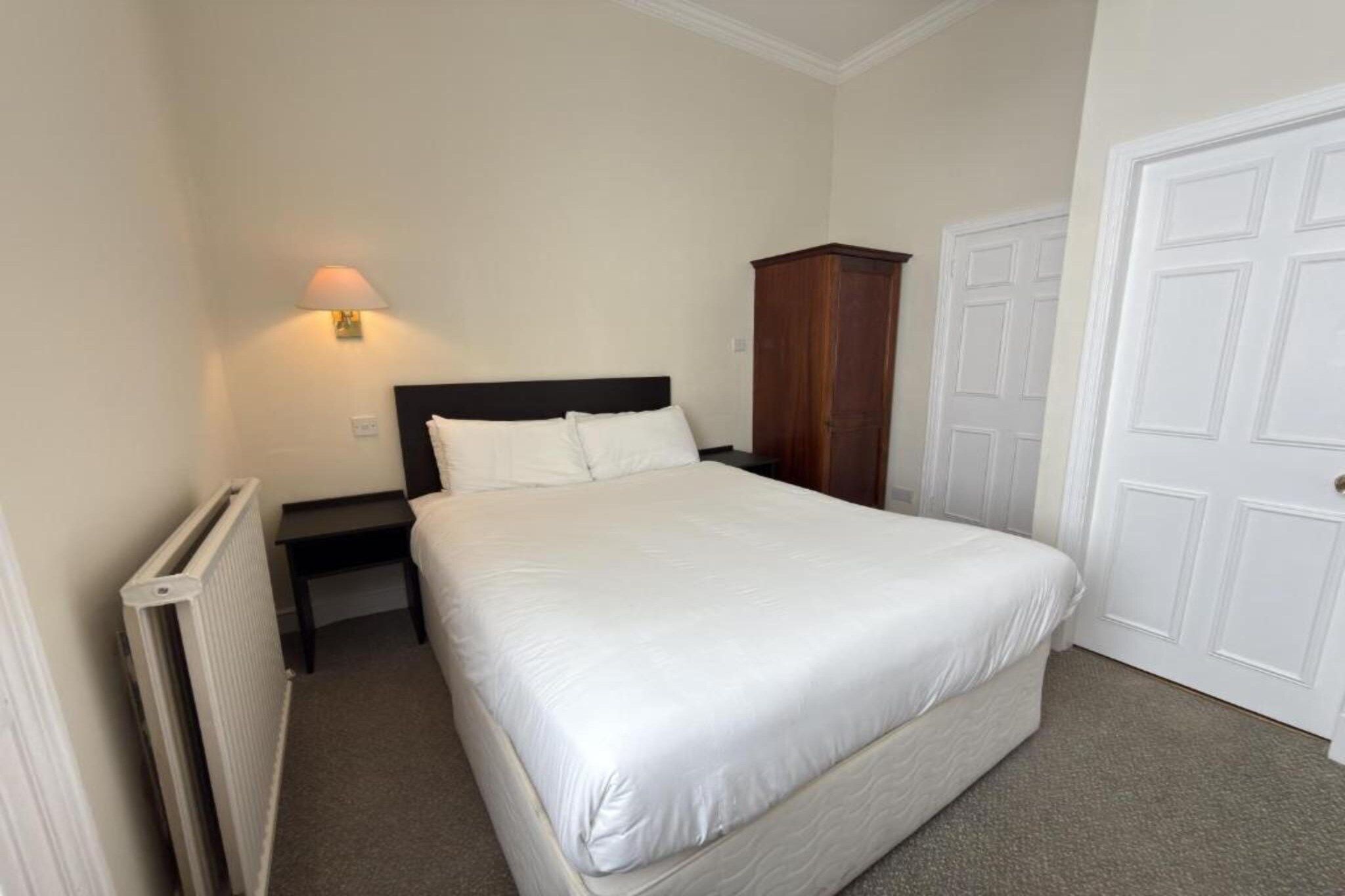 A bedroom with a bed, a nightstand, a wall-mounted lamp, a wardrobe, and a door.