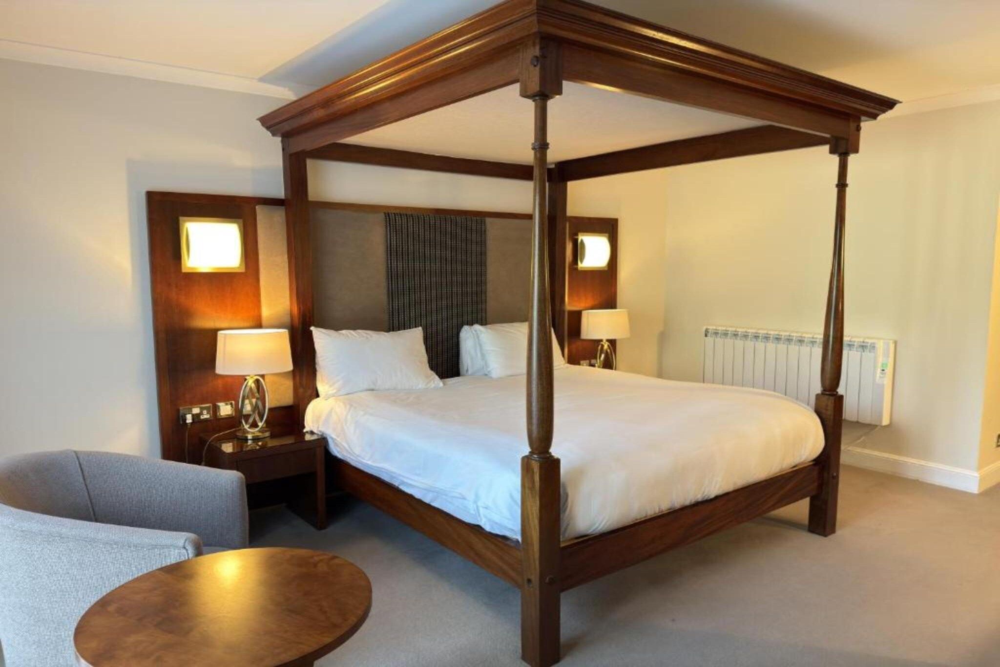 A four-poster bed in a hotel room with a nightstand, lamp, and a round table.