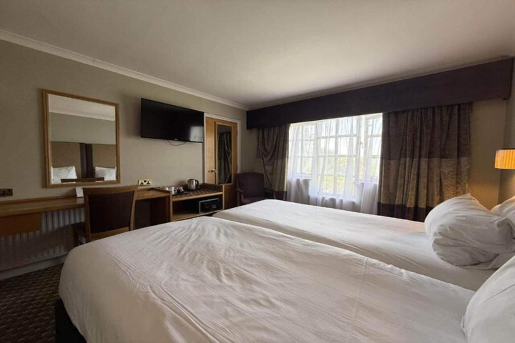 Une chambre d’hôtel avec deux lits, un bureau, une chaise, une télévision et une fenêtre avec des rideaux.