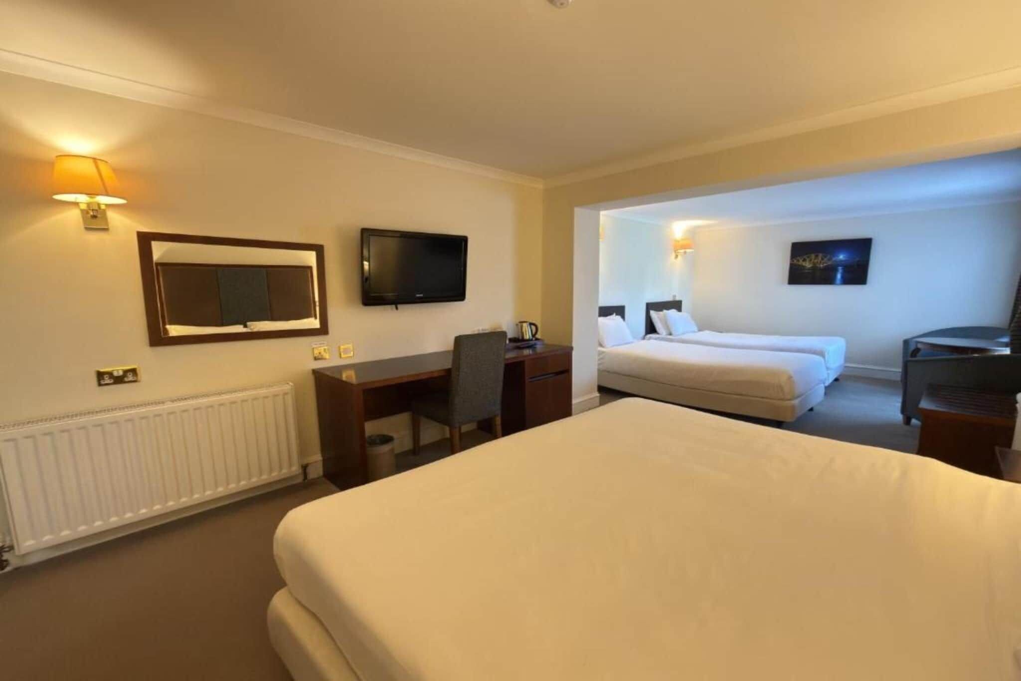 Habitación de hotel con una cama grande, un escritorio, una silla, un televisor y una cama con dos almohadas.
