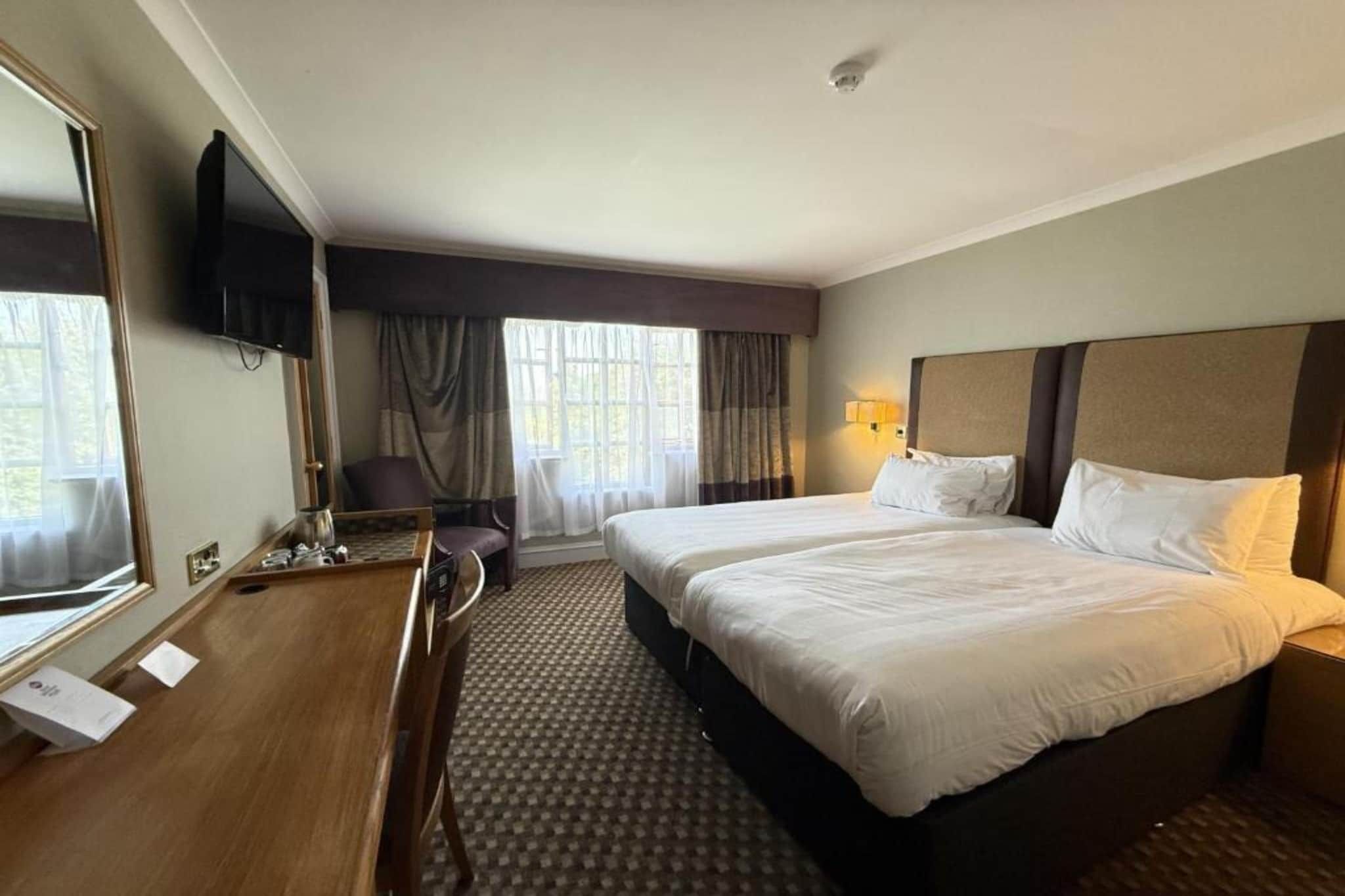 Habitación de hotel con cama, escritorio, silla, televisor y ventana con cortinas.