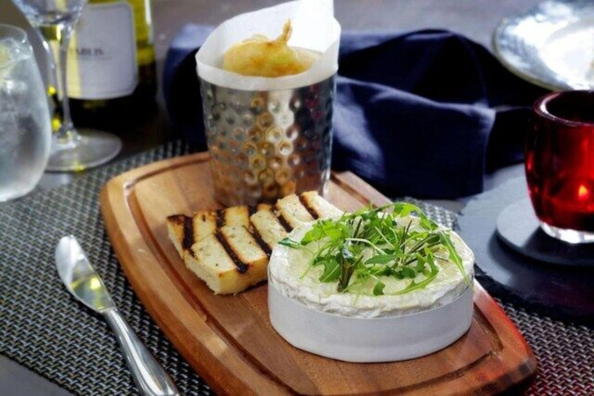 A round cheese wheel garnished with greens, accompanied by toasted bread slices and a small container of sauce on a wooden board.