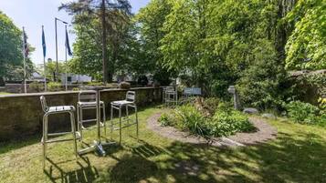 Een tuin met metalen stoelen, een stenen muur en diverse bomen.