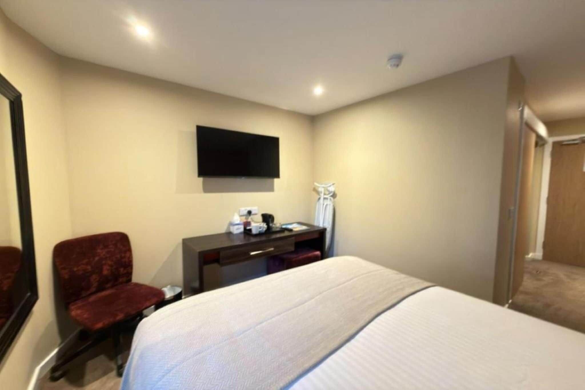 Ein Hotelzimmer mit Bett, Schreibtisch, Stuhl, einem an der Wand befestigten Fernseher und einer Tür.