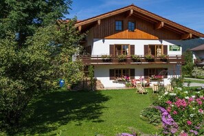 A two-story wooden chalet with a balcony, surrounded by a well-maintained garden and trees. - Holiday apartment with 1 bedroom (Inzell)