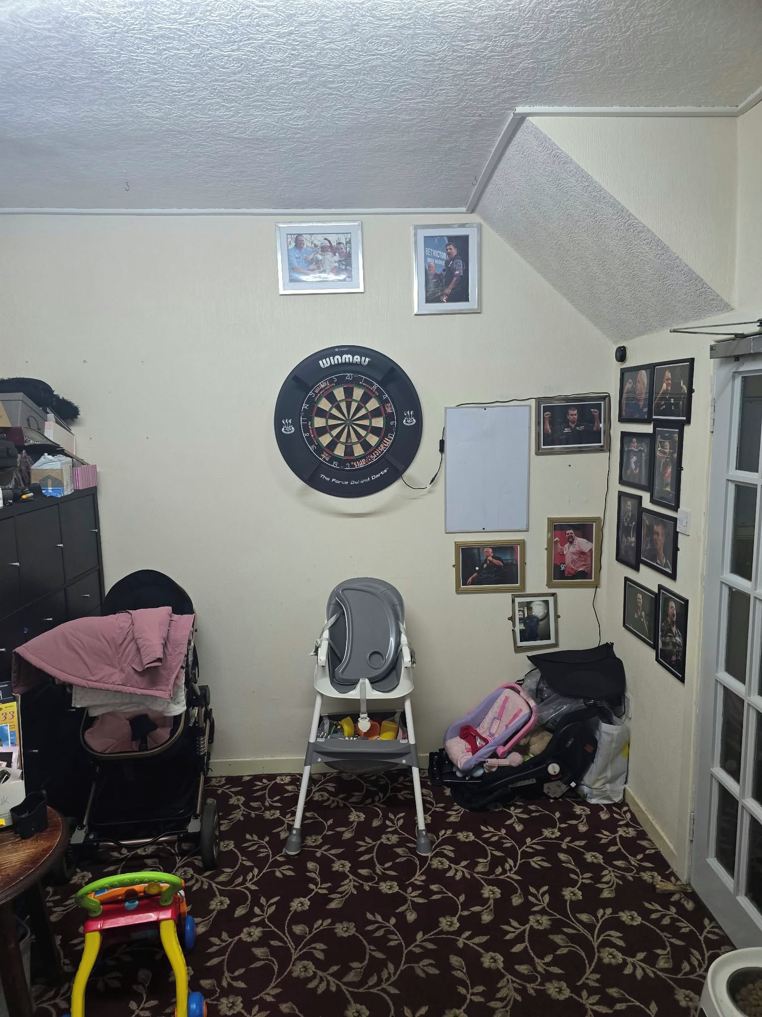 Ein Zimmer mit Dartscheibe, Kinderwagen, Rollator und einer Tür mit Fenster.