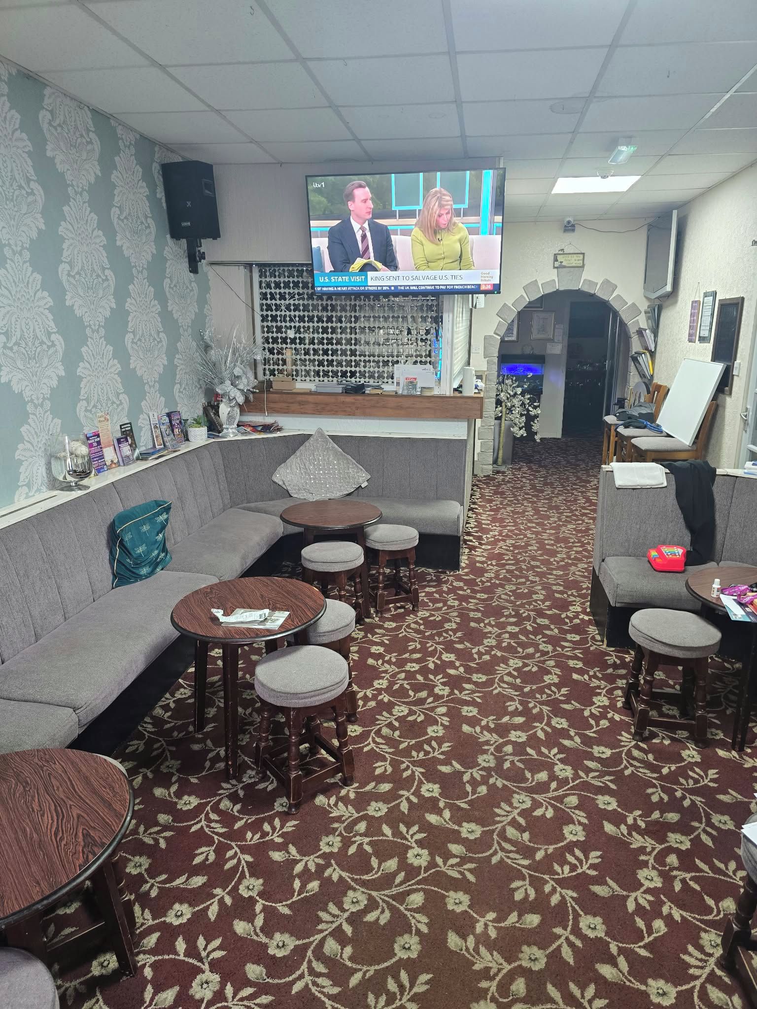 Eine Hotelhalle mit Fernseher, der Nachrichten zeigt, einem Empfangsbereich, einer Sitzecke und einem gemusterten Teppich.